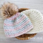 Two simple crochet hats laying in a basket on a table. One hat is colorful with a pom pom and the other is a neutral cream color.